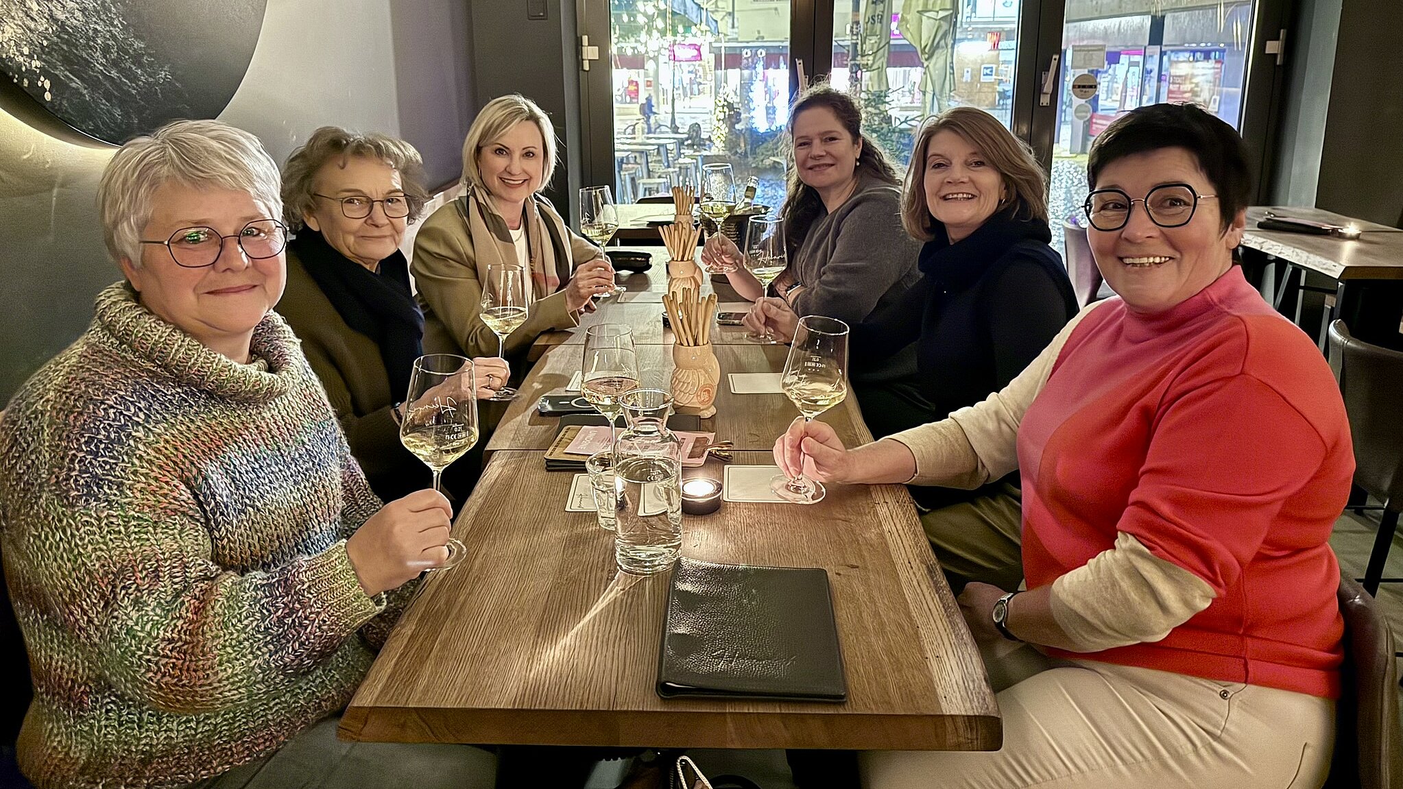 Sechs Frauen sitzen an einem Tisch in der Weinbar und lächeln in die Kamera