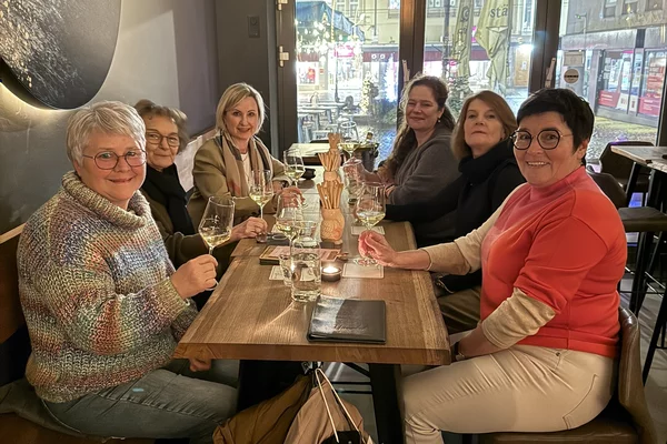 Sechs Frauen sitzen an einem Tisch in einer Weinbar und lächeln in die Kamera